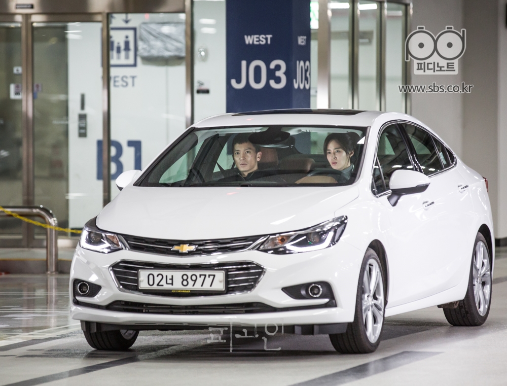 흰색 차 안에 지성과 권유리가 타 있고, 배경은 지하 주차장이다