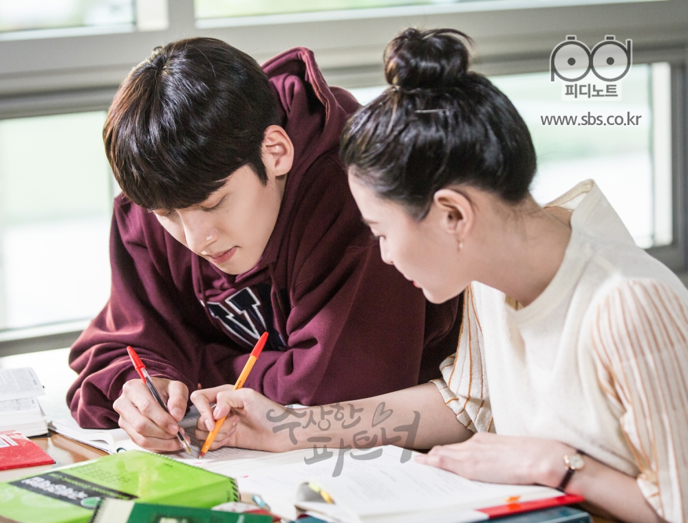 지욱과 유정이 대학교 도서관에서 나란히 앉아 공책에 함께 낙서하는 측면의 모습.