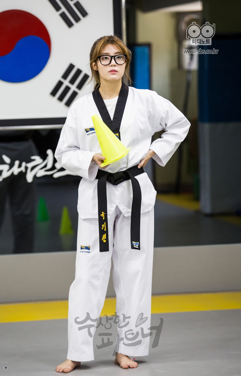 태권도장에서 도복을 입고 뿔테 안경을 쓴 봉희가 노란색 출석부를 들고 서있는 봉희.